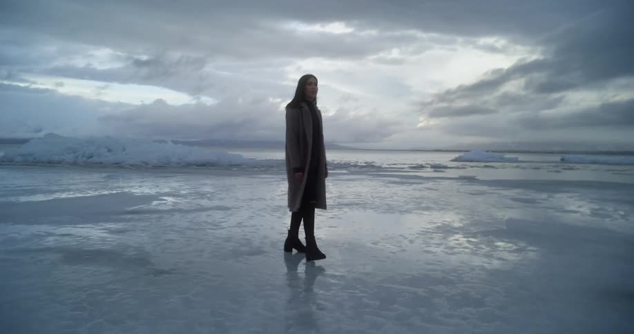 A woman in a long winter coat walking alone over snow and ice