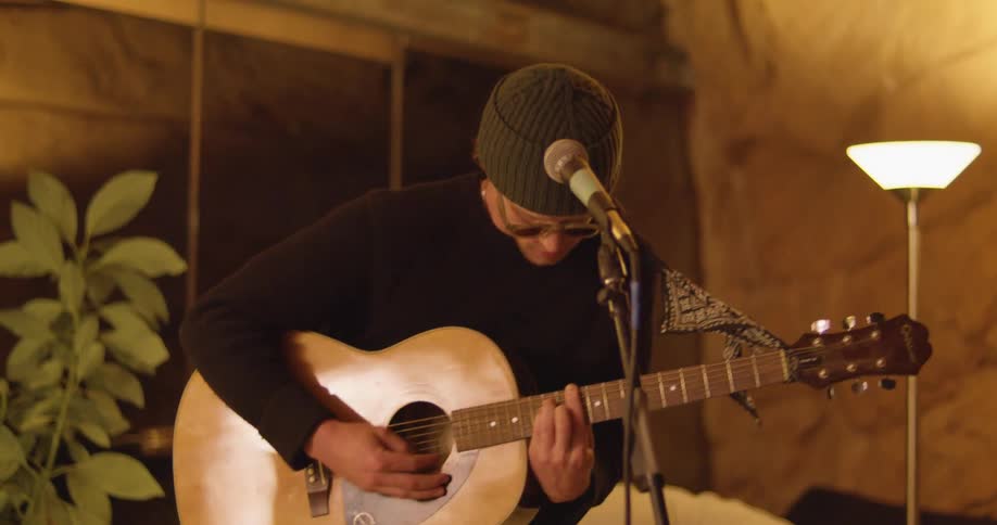 Man wearing knit hat and sunglasses plays guitar standing next to microphone