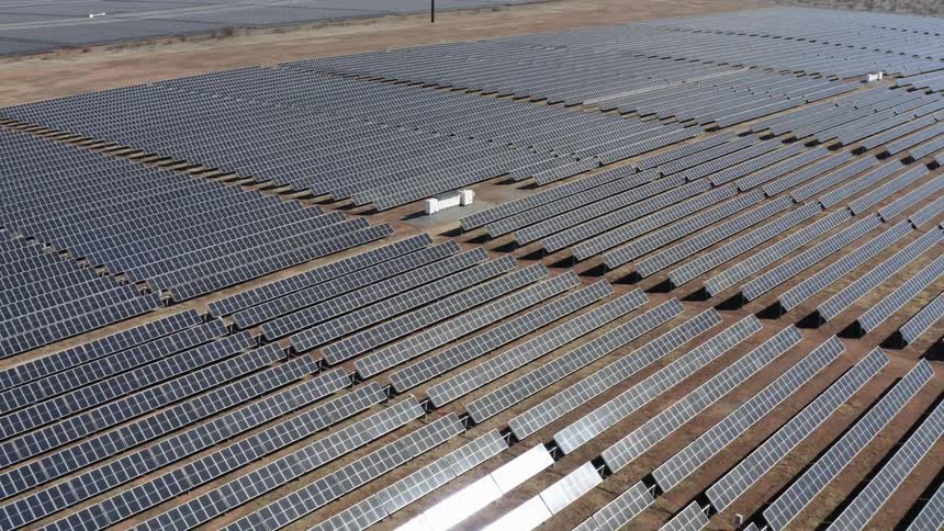 Aerial view of rows of black solar panels reflecting sunlight on a dirt field