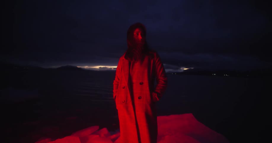 A woman with a red light shining on her standing up outdoors with her hands in her pockets looks at the camera and then looks away