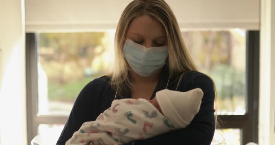 A woman wears a face mask as she cradles and rocks her newborn while standing in front of a large window