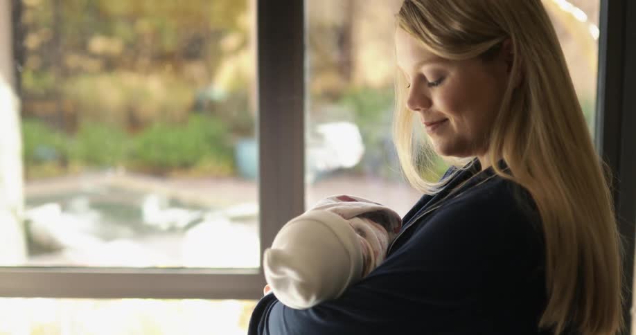 A woman holding a baby with a blanket and hat with a window in the background