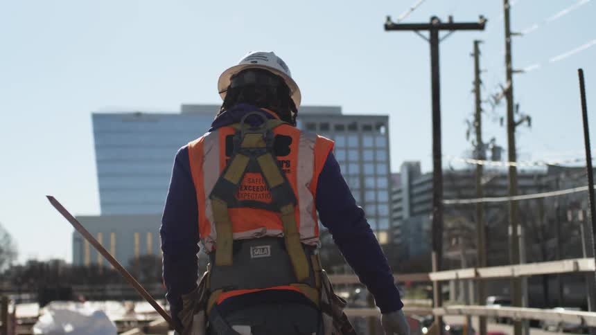 A person with gloves and a hardhat walking with buildings the sky and clouds in the background