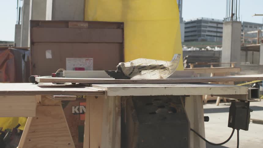 A person with gloves cutting a piece of wood with an electrical saw with buildings the sky and clouds in the background