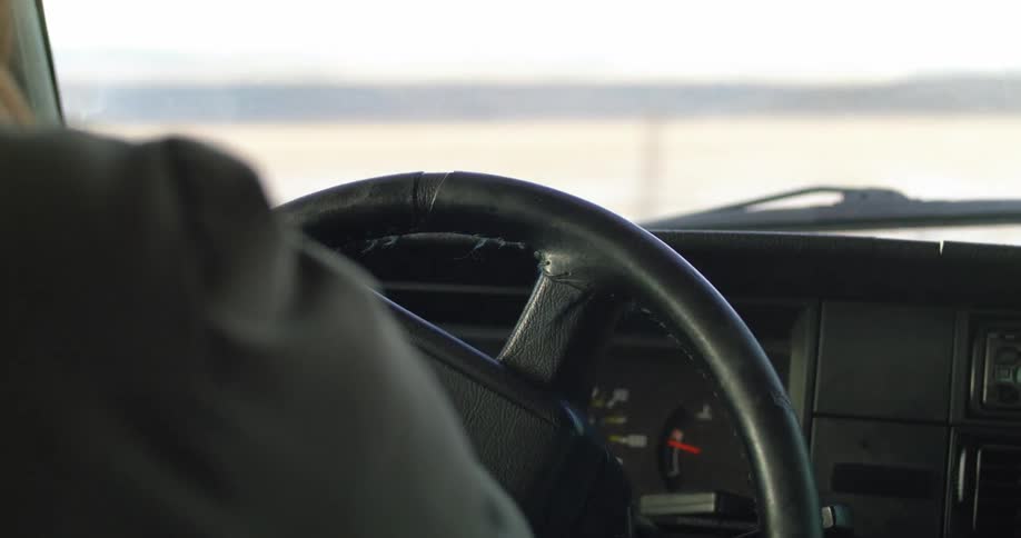 A woman driving a vehicle and turning the steering wheel with a landscape the sky and clouds in the background