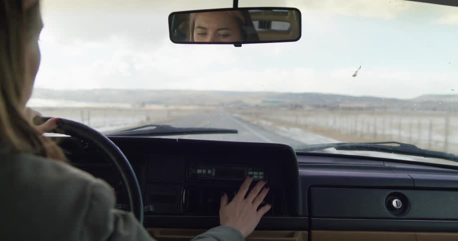 A woman holds her hand over the air vent in a vehicle as she drives down a countryside road during the day