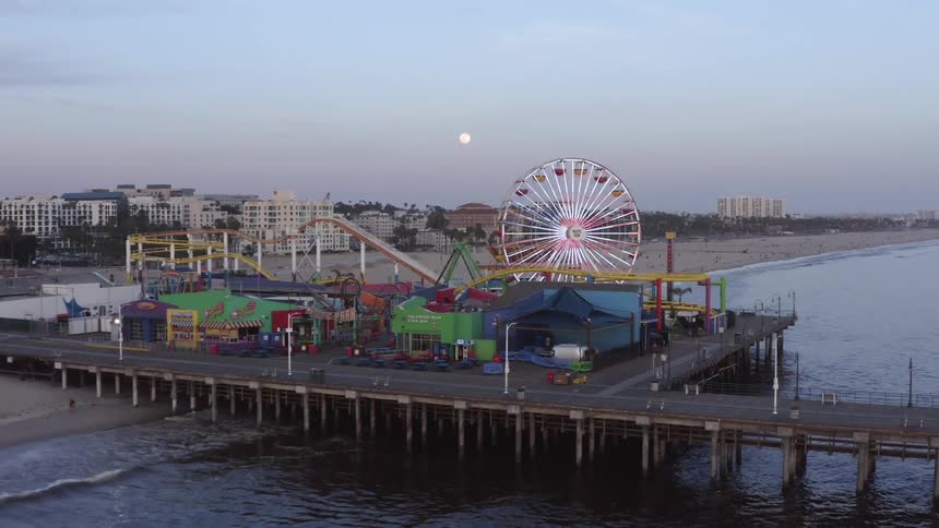 A beach with a boardwalk and a ferris wheel with buildings the sky the moon and clouds in the background