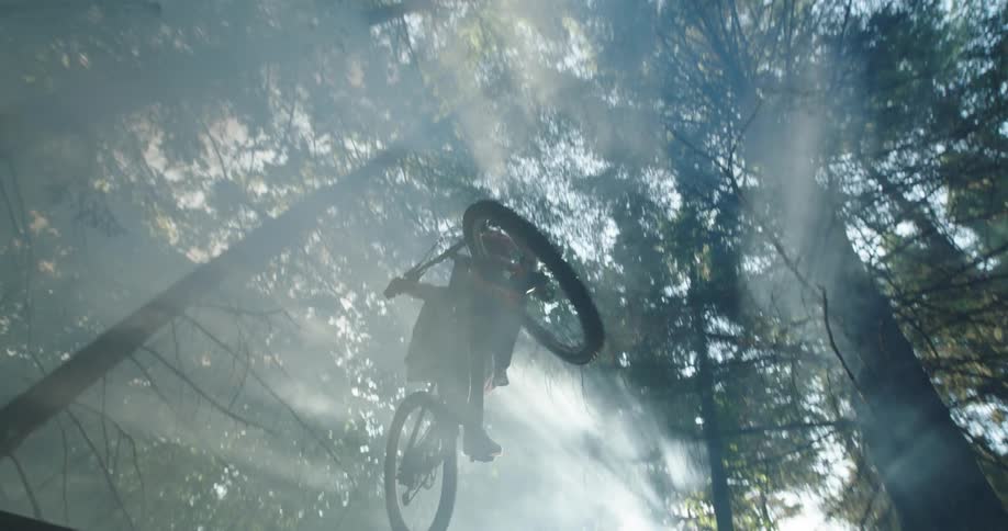 Sunlight filtering through the fog in the forest lights the silhouette of a man jumping a mountain bicycle through the air