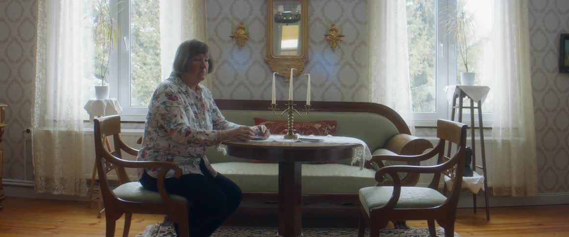 An elderly woman sits alone at a small table where she takes off her reading glasses