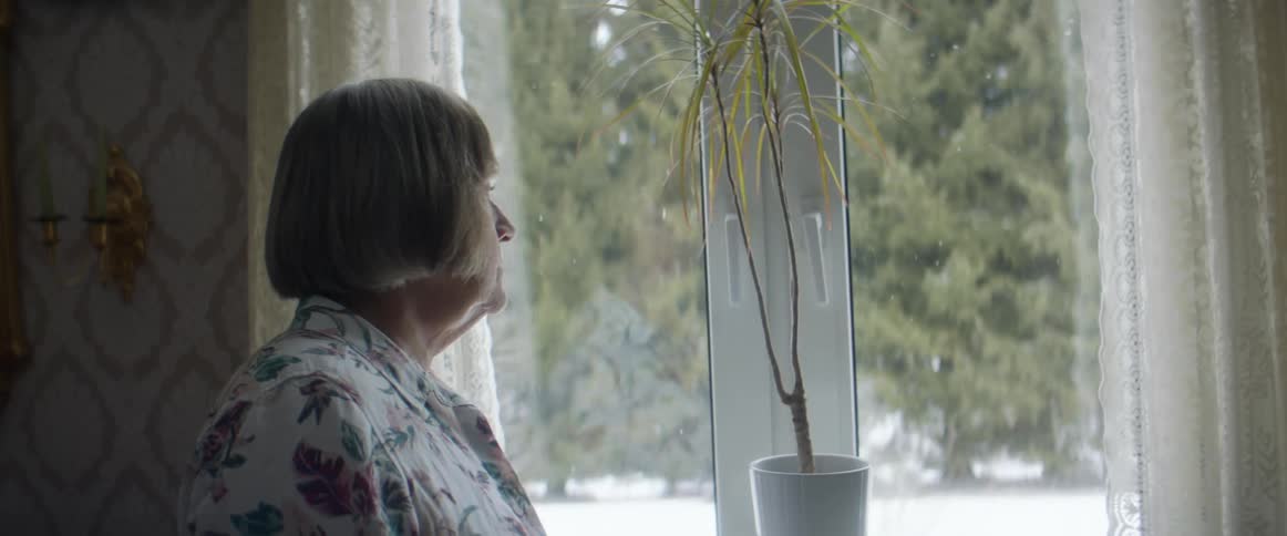 A woman standing in a room stares out of a window with a sad look on her face at daytime