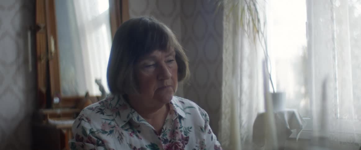 An old woman sitting in a room at home takes her glasses off and stares off sadly