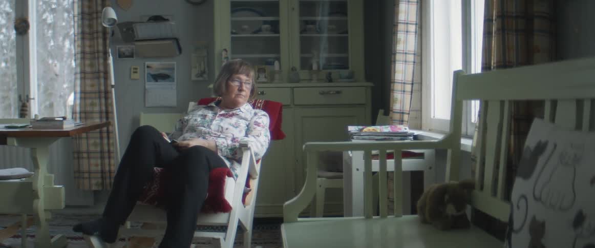 A woman sits in a rocking chair in her home and looks out the window in disappointment
