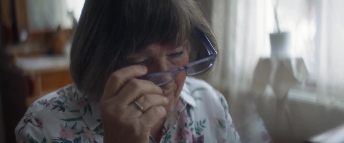 An elderly woman sitting at a table takes off her glasses while looking a bit bored