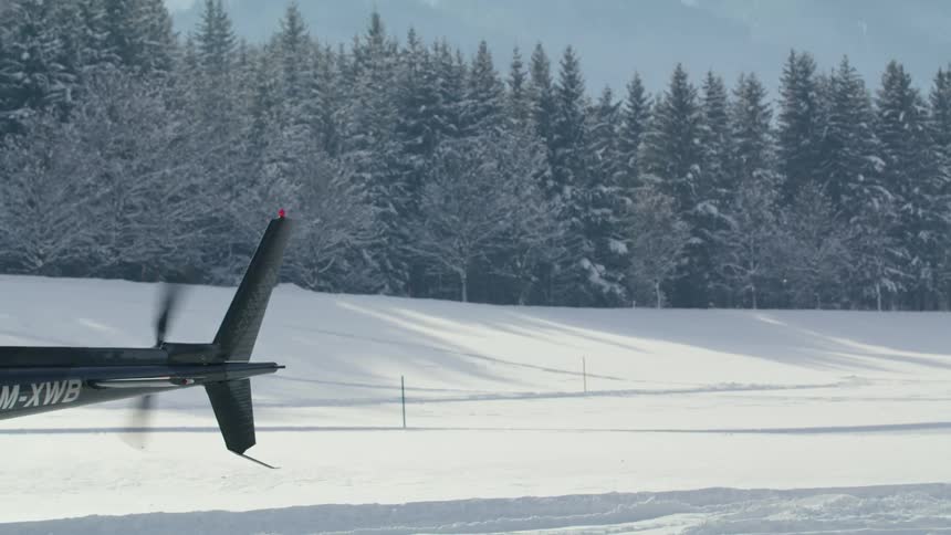 Helicopter propellers spin as it sits on the ground with snow covered trees in the distance