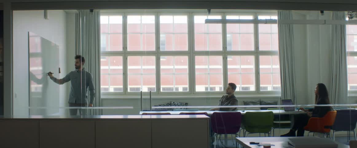 A man with a marker writing on a whiteboard with another man and a woman sitting on chairs at a conference room table