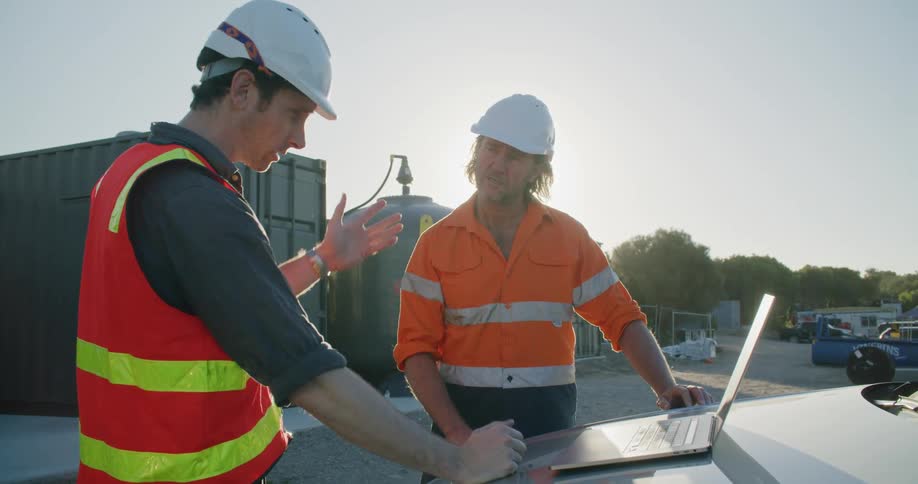 Two men with hardhats looking at a laptop with trees the sky and clouds in the background