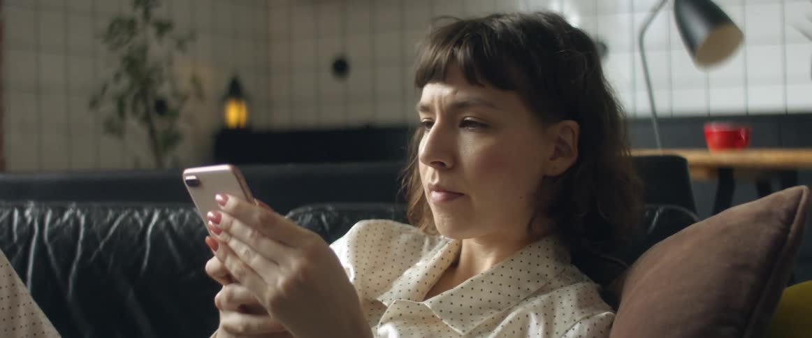 A woman wearing pajamas puts a thermometer under her arm to take her temperature then picks up her phone