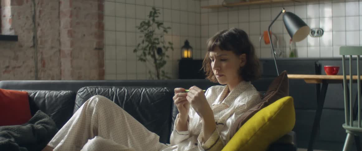 Woman sitting in a couch at home takes her temperature and checks her smartphone