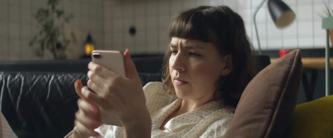 A woman sits on a couch looking at her phone then crosses her arms in frustration