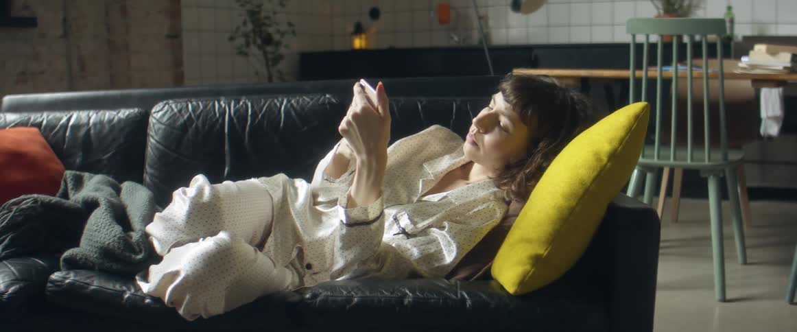 A woman lays on her couch in her pajamas while looking at her smart phone