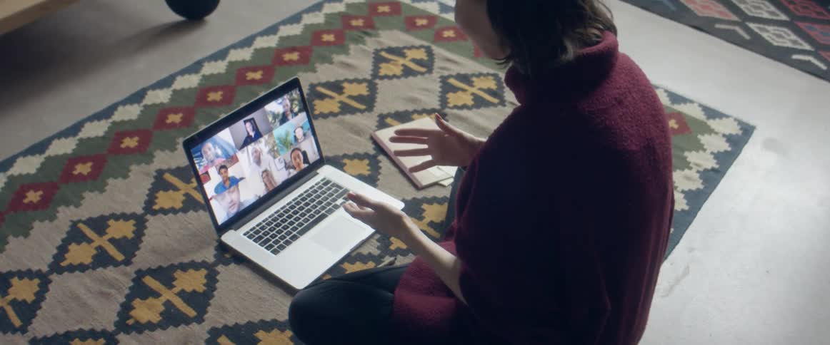 A woman sitting on a rug on the floor talks to a screen full of people on her laptop