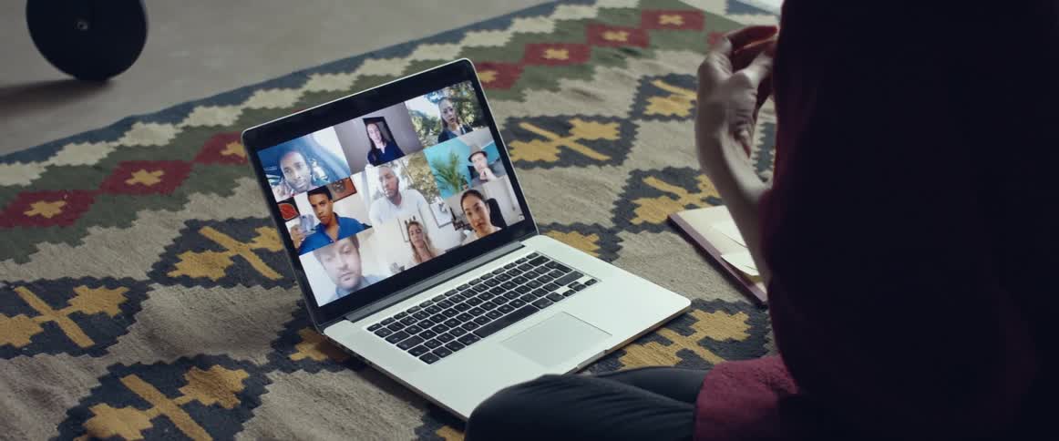 A person with a pen sitting on the floor on a carpet in front of a laptop