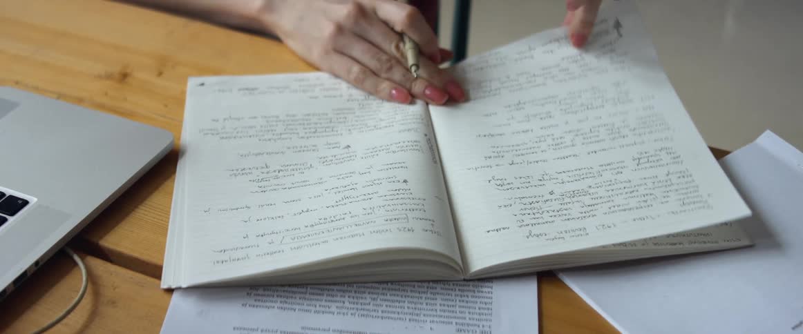 A woman sitting on a chair at a desk with a laptop looking through a book