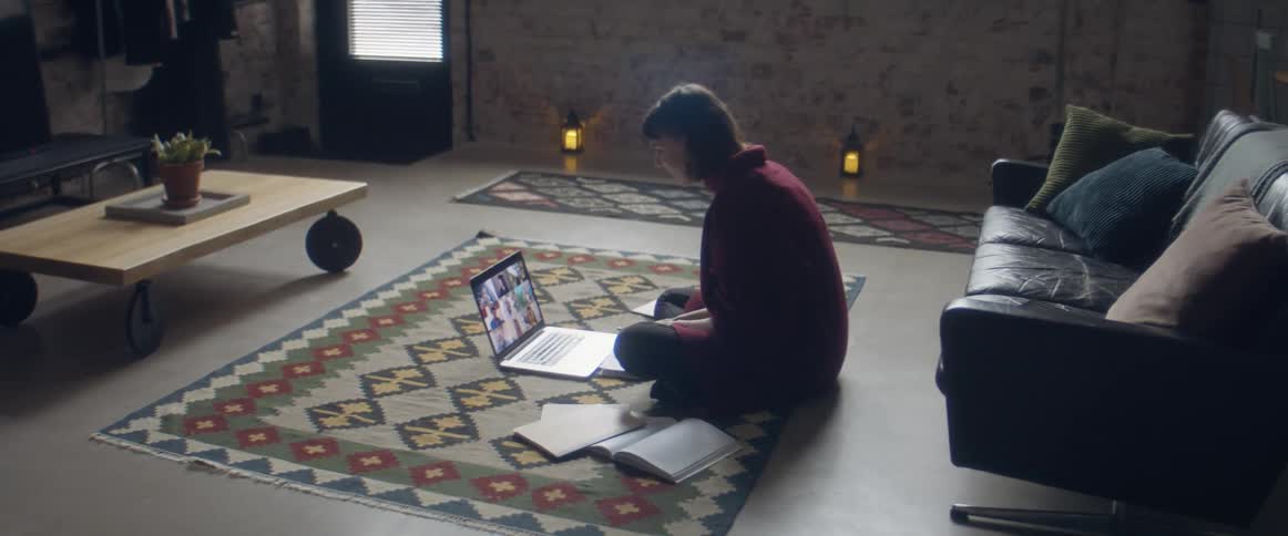 A teenage girl sits on the floor at home and video chats on a laptop