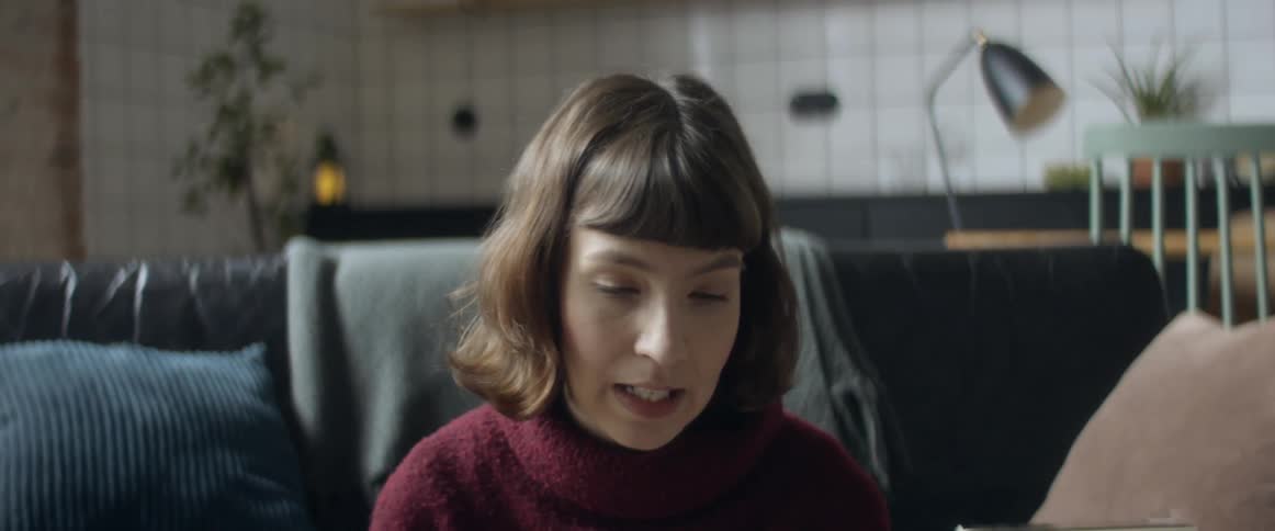 Woman wearing sweater talks sitting on couch near white tiled kitchen