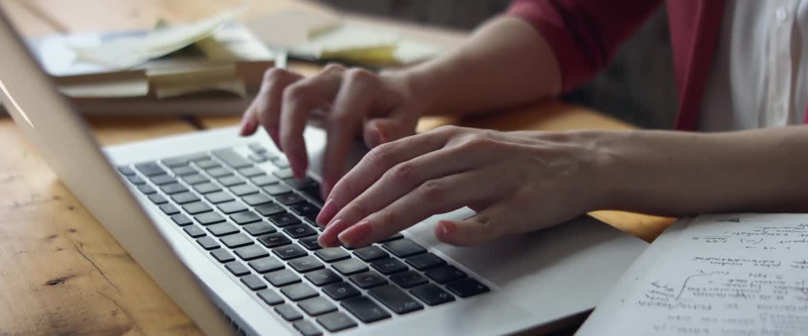A woman sitting at a wooden table types on the keyboard of her laptop computer