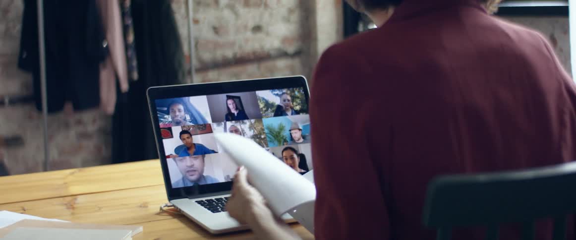 A woman sits at a table having a video call on a laptop and looking through a stack of papers