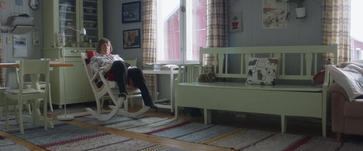 An older woman sits alone in a rocking chair and takes off her glasses