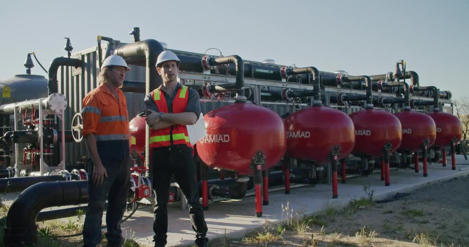 Two men with hardhats standing with pipes the sky and clouds in the background