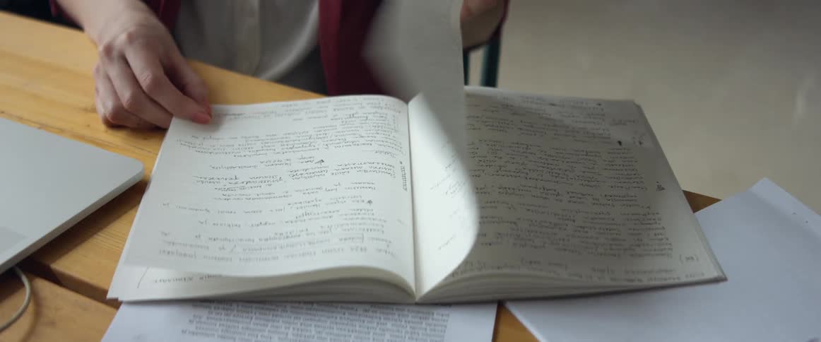 A woman flips through a notebook before turning to type on a laptop while sitting at a desk