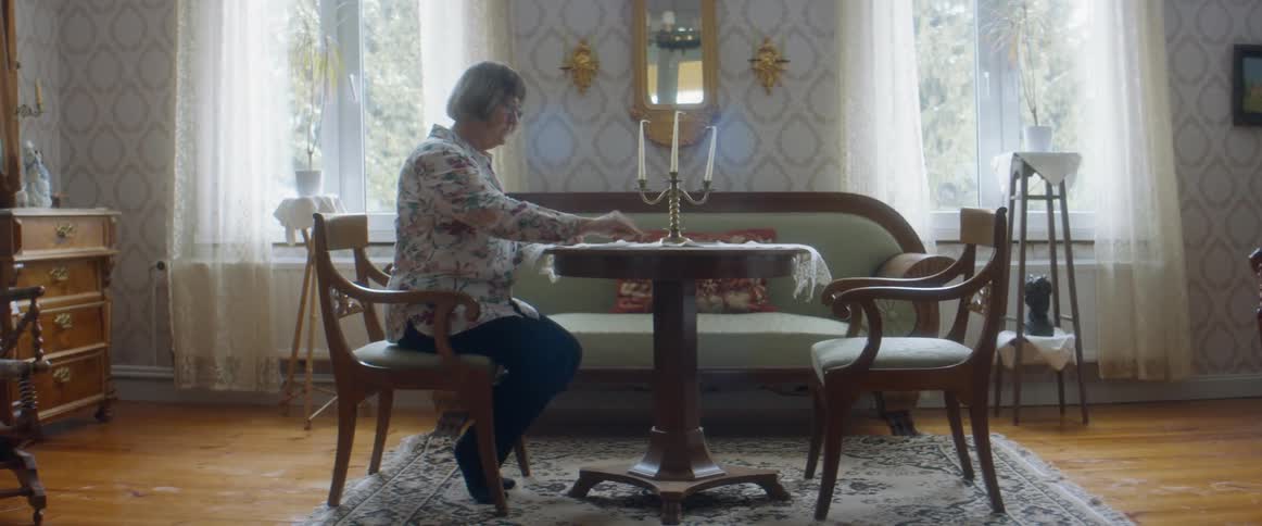 An elderly woman sits at a small table in her home while laying out playing cards to play solitaire