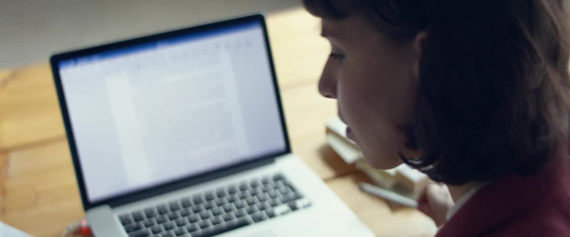 Woman reviews papers before typing on laptop sitting on wooden plank table