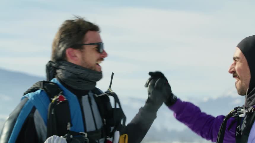 Two men high five and clasp hands standing outdoors with snowy mountains in background
