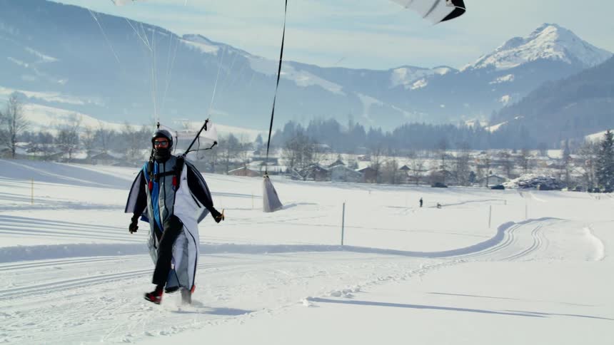Parasailing man lands on snowy plain with town and mountains in background as person stands and watches