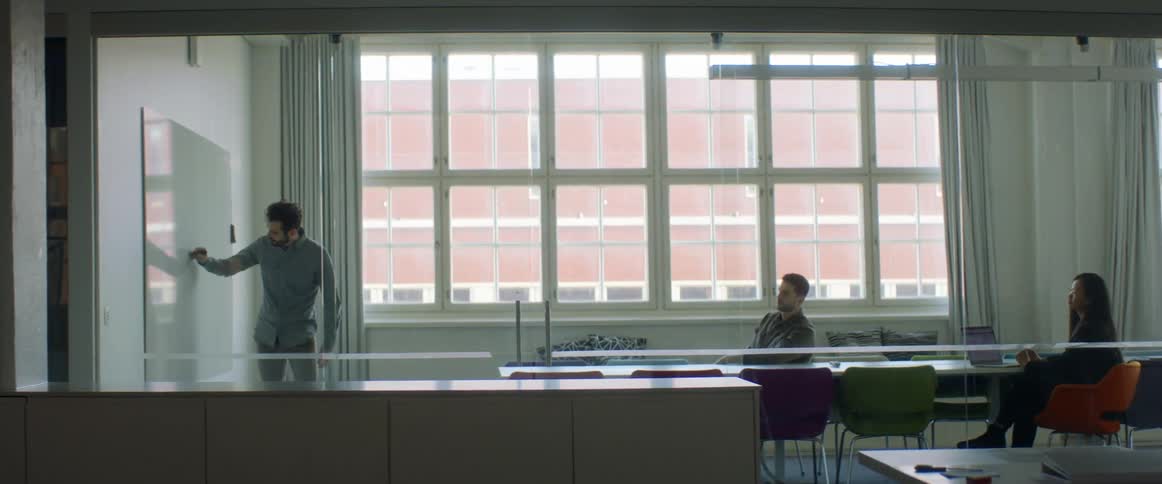 A man standing in a room writing on a whiteboard with another man and woman sitting on chairs by a conference table