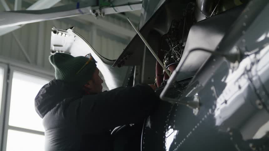 A man with a coat and hat working on a aircraft with a wall and window in the background