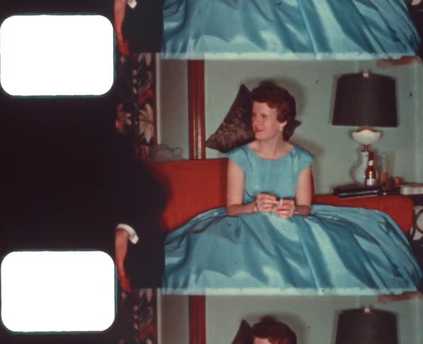 A woman in a blue dress sitting on a couch next to a man with a wall and a lamp in the background