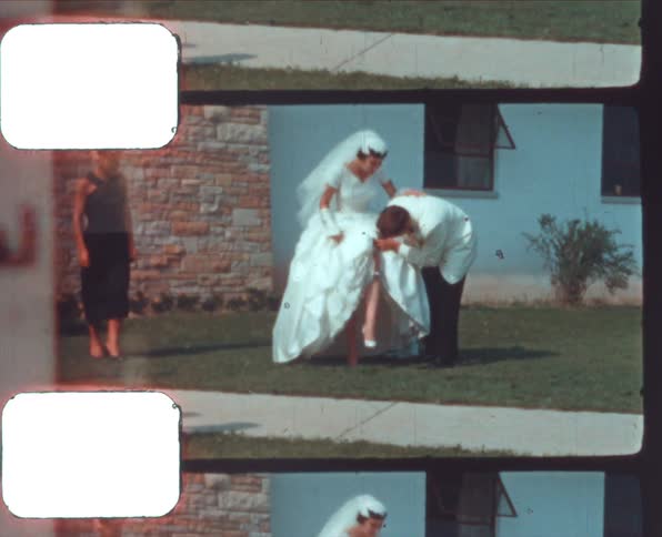 A woman stands by watching while a groom helps his bride remove her wedding garter outdoors in front of a building at daytime