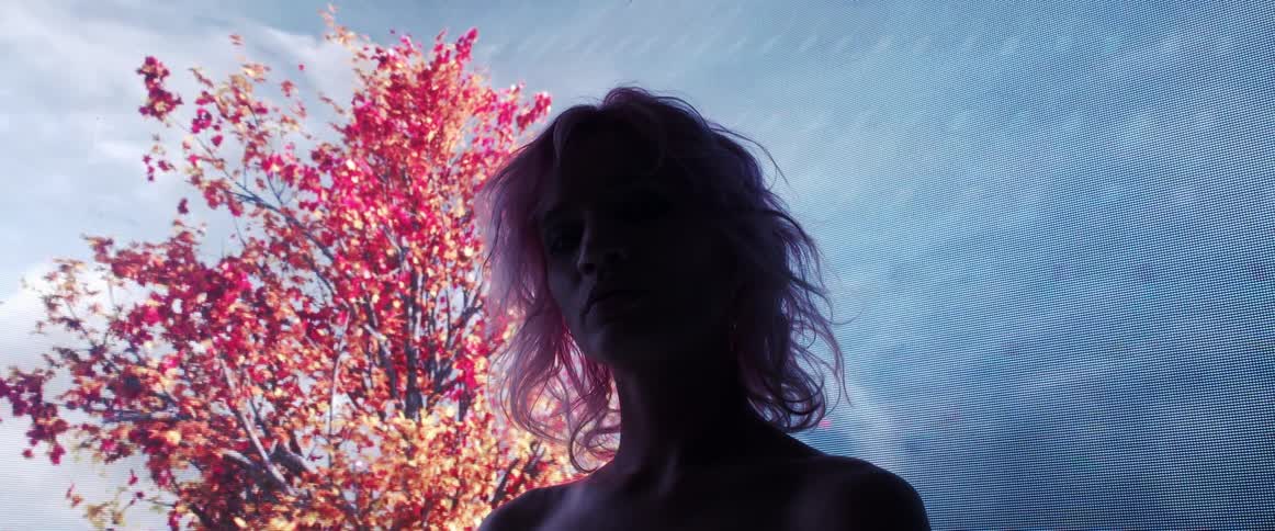 The silhouette of a woman is illuminated by a large screen displaying the sky and trees behind her