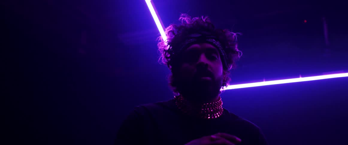 A man wearing a thick gold chain necklace and a hair bandana poses under neon lines