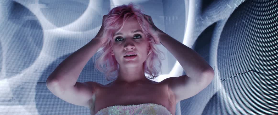 A woman with pink hair looking at the camera in a room with circular digital images moving across the walls