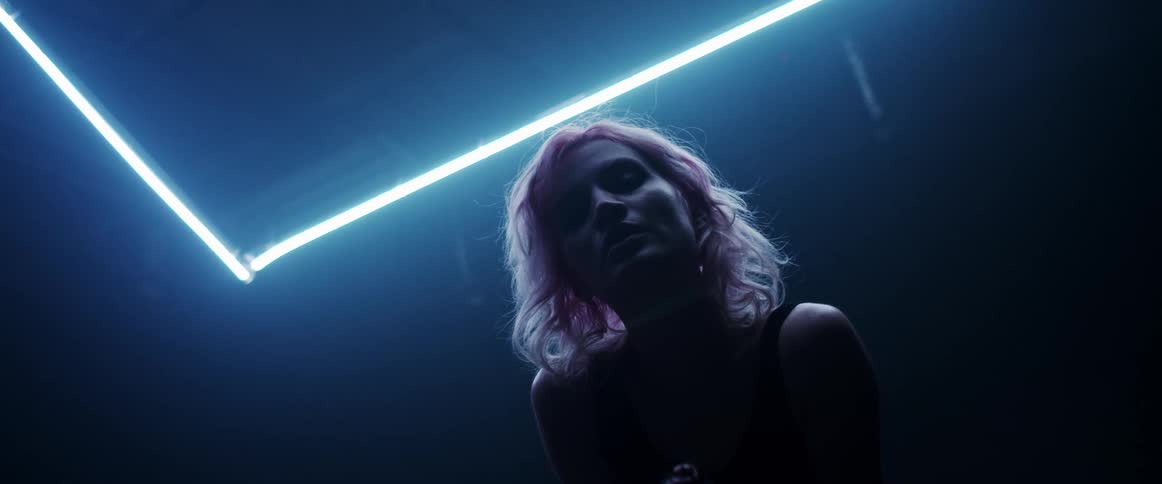 A woman with short blond hair looks down at the camera while a neon light above illuminates her