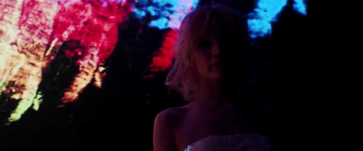A woman stares into the camera while standing in front of a video wall in a dark room
