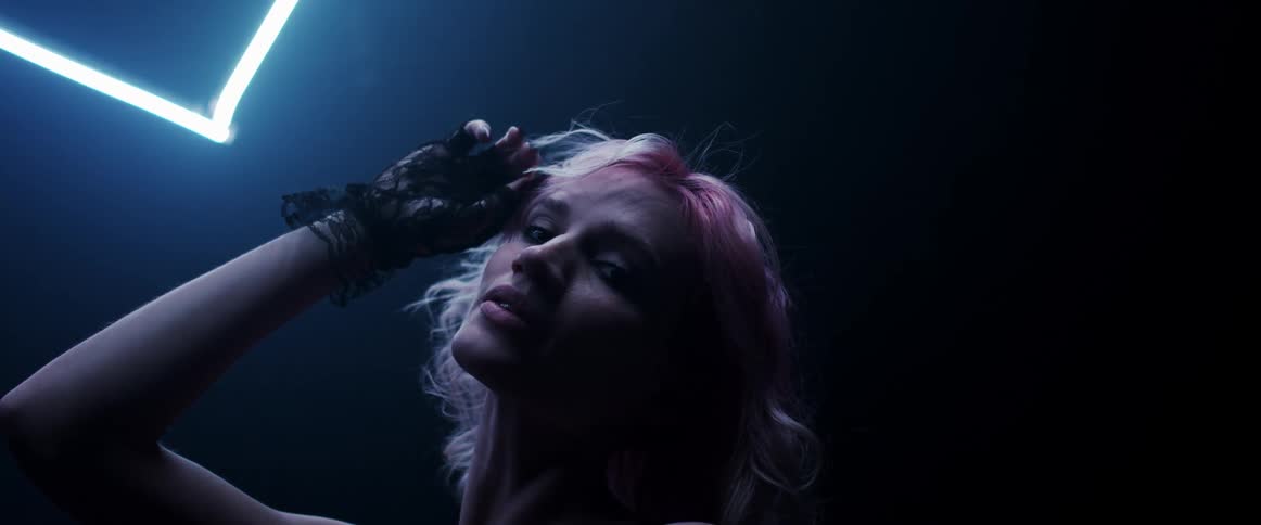 A woman with short blonde hair stands in a darkened room and subtly smiles at the camera