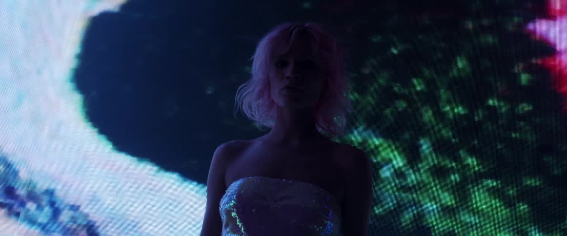 A model in a sequined dress poses in front of a large screen showing digital graphics