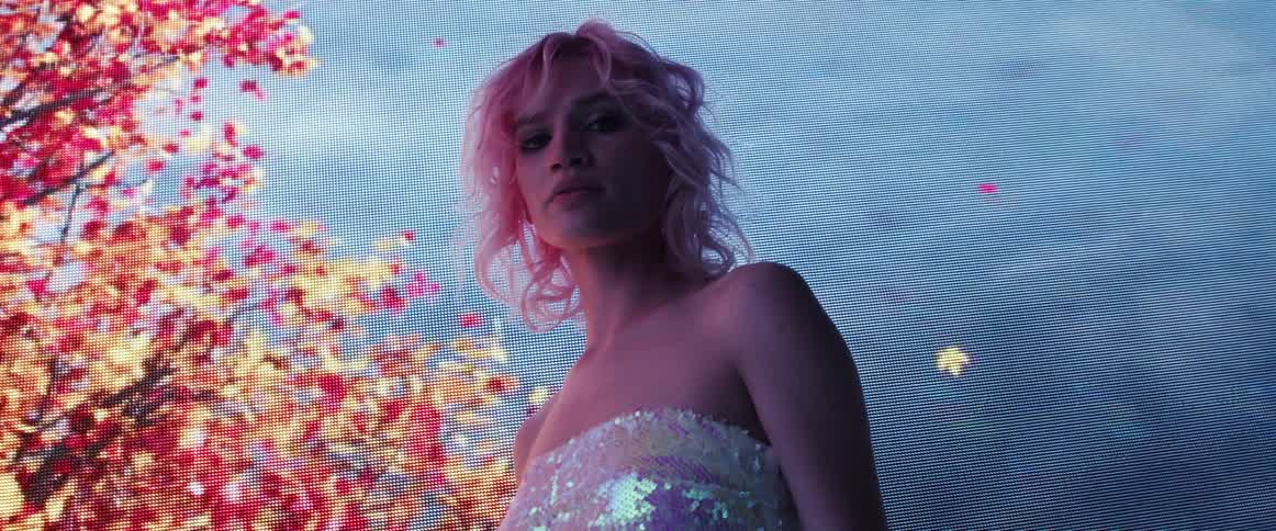 A model with pink hair poses in front of a digital screen with images of flowers blooming on a tree